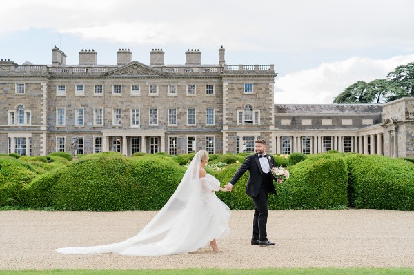 Bride and groom walk along pathway to wedding venue castle