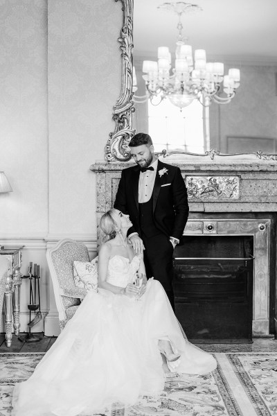 Black and white image of bride fixing her earrings sitting down groom behind her