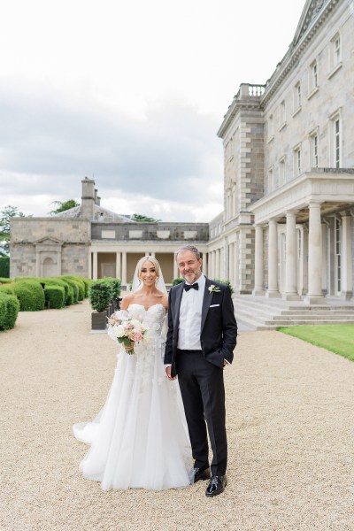 Father of the bride poses outside wedding venue she holds bouquet