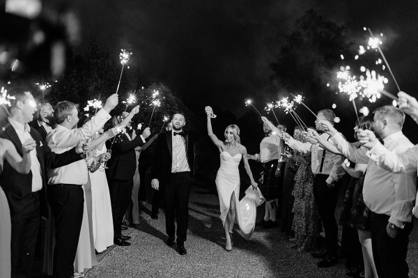 Bride and groom walk through line of sparklers held up by guests black and white