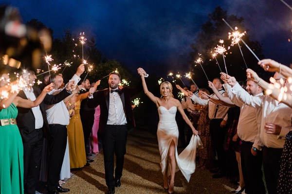 Bride and groom walk through line of sparklers held up by guests