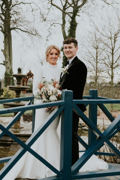 Bride and groom stand on bridge in garden