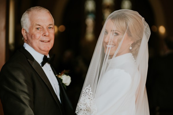 Brides face is covered with veil she stands beside her father