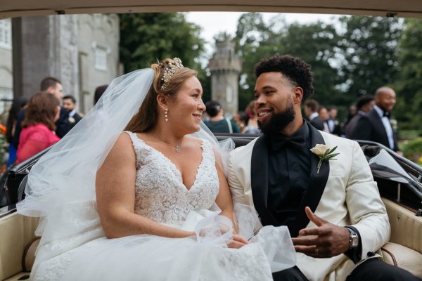 Bride and groom sit smiling at each other in wedding car