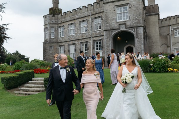 Mother father and bride walk on the grass outside wedding venue