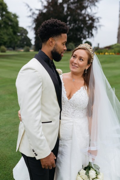 Bride and groom stand on grass together smile at each other