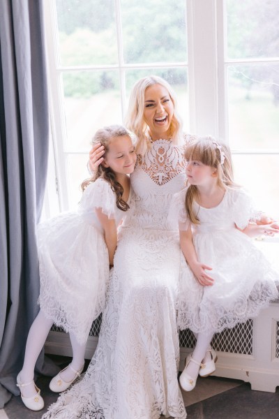 Bride and two daughters wearing white dresses stand at windowsill