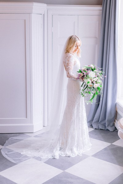 Bride stands ready for wedding facing window holding bouquet of flowers