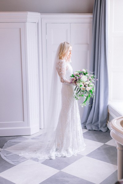 Bride stands ready for wedding facing window holding bouquet of flowers