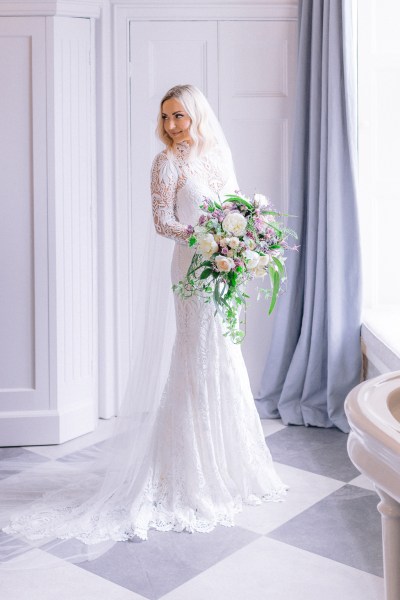 Bride stands ready for wedding facing window holding bouquet of flowers