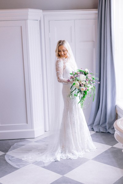 Bride stands ready for wedding facing window holding bouquet of flowers