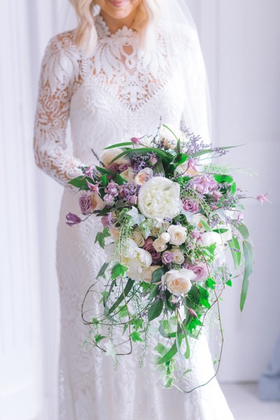 Bride stands ready for wedding facing window holding bouquet of flowers