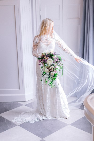 Bride stands ready for wedding facing window holding bouquet of flowers showing off veil detail