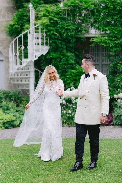 Bride and groom stand in garden on the grass together staircase behind them