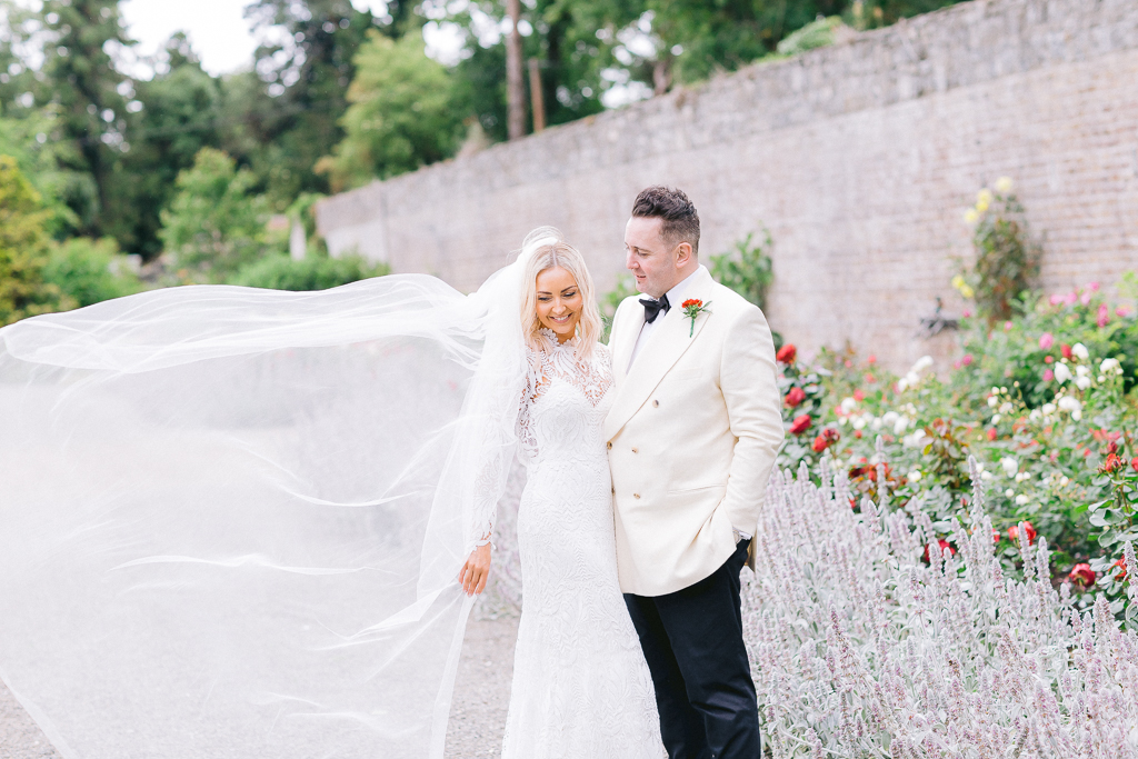 Brides veil blowing in the wind groom looking at her in garden