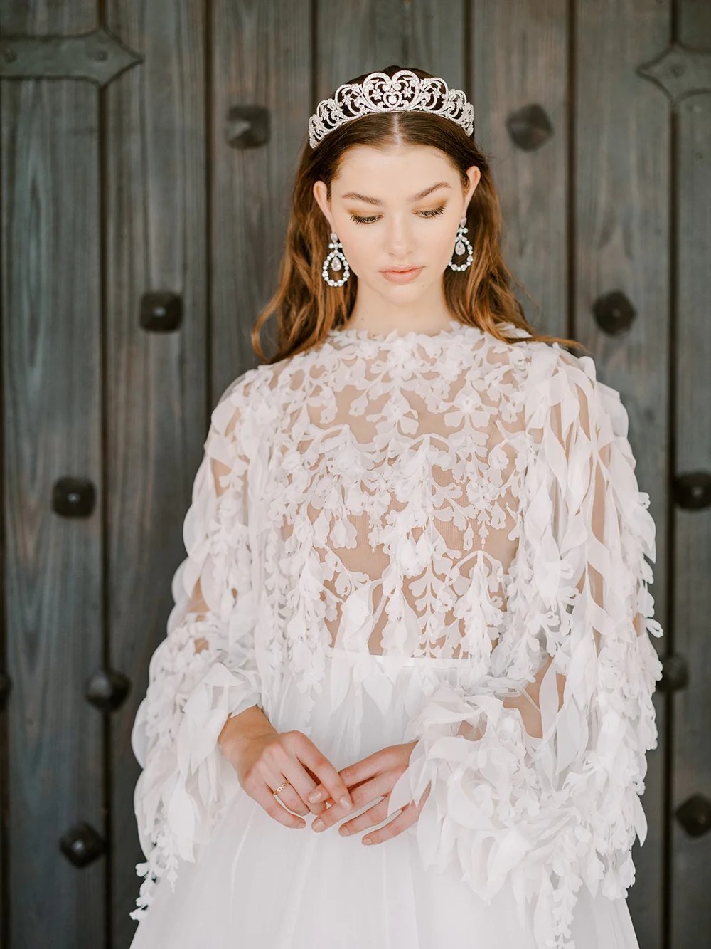 A model wears an Eden Luxe tiara with a lace and feather sheer dress standing against a door