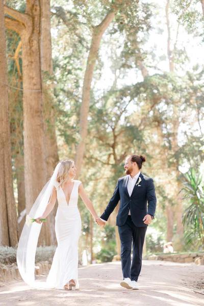 Bride and groom walk hand in hand along pathway in park forest setting