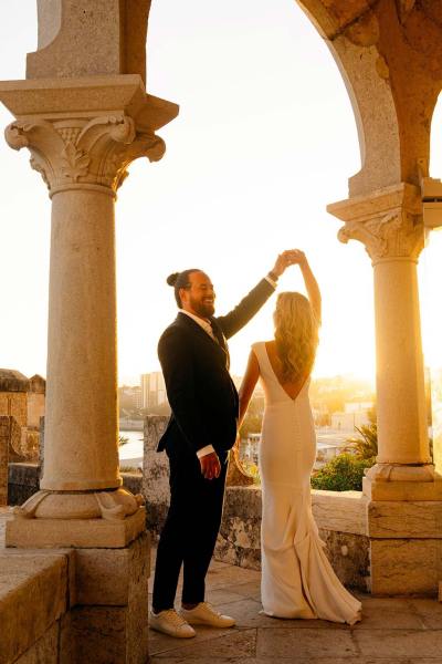 Bride and groom stand in front of pillars sunset in background twirl dancing on balcony