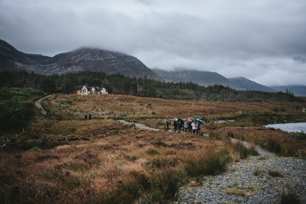 Trail of guests walking uphill on pathway holding black umbrellas mountain in background