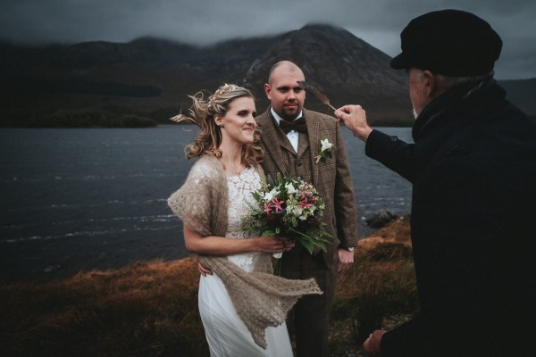 Bride and groom stand smiling in front of celebrant he holds a feather to their faces