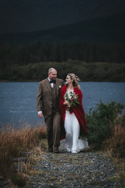 Bride and groom walk along pathway to edge of cliff bride wears red shawl