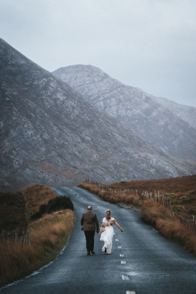 Bride and groom walk hand in hand along dirt road mountains in background scenery