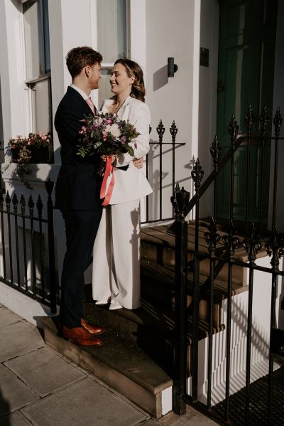 Bride and groom walk hand in hand together in the sun residential road she holds the bouquet in hand they embrace on steps