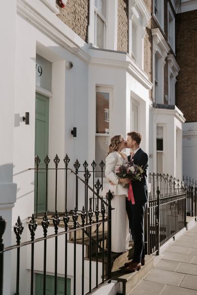 Bride and groom walk hand in hand together in the sun residential road she holds the bouquet in hand they kiss