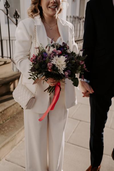 Bride and groom walk hand in hand together in the sun residential road she holds the bouquet in hand