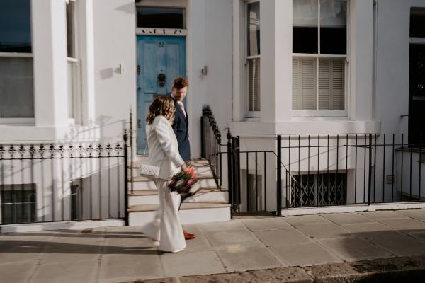 Bride and groom walk hand in hand together in the sun residential road she holds the bouquet in hand