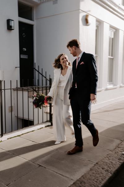 Bride and groom walk hand in hand together in the sun residential road she holds the bouquet in hand