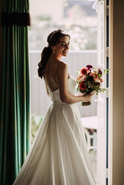 Bride stands at the window holding bouquet of flowers windowsill and curtains smiling