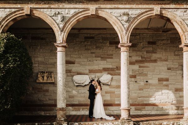 Wideshot of bride and groom standing under pillar archway holding each other