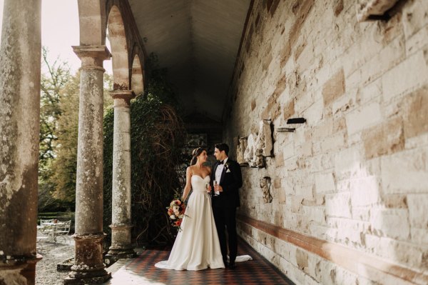 Bride and groom look at each other stand in front of bricked wall holding glasses of prosecco wideshot