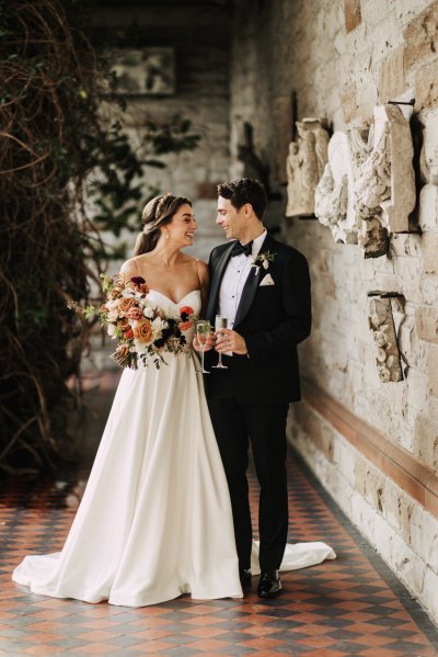 Bride and groom look at each other stand in front of bricked wall holding glasses of prosecco