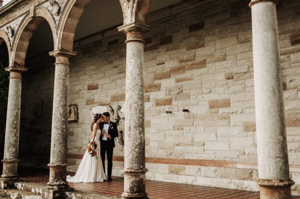 Bride and groom kiss and embrace under archway pillars cathedral