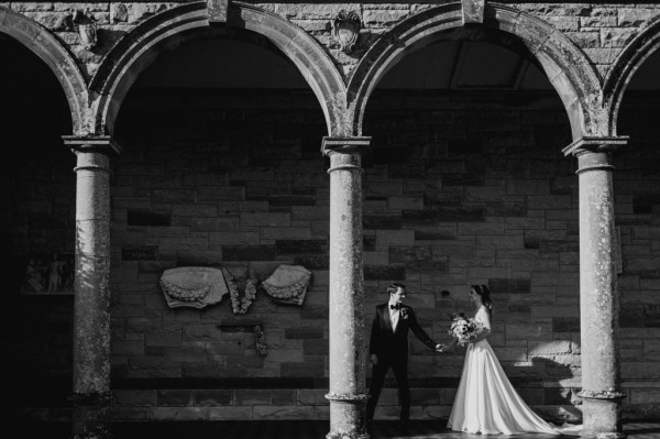 Black and white shot of bride and groom standing under pillar archway