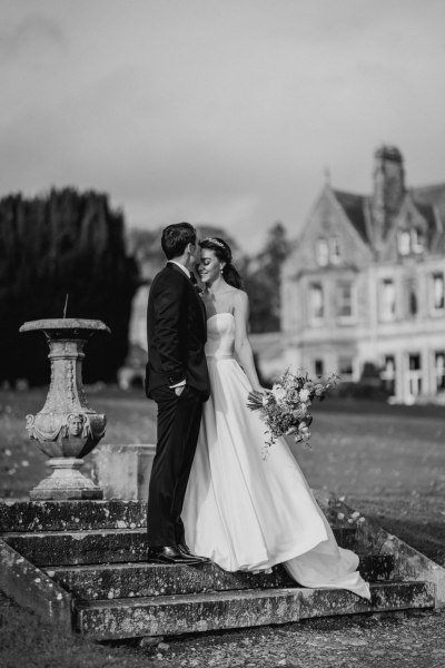 Black and white image of bride and groom in front of venue standing on steps