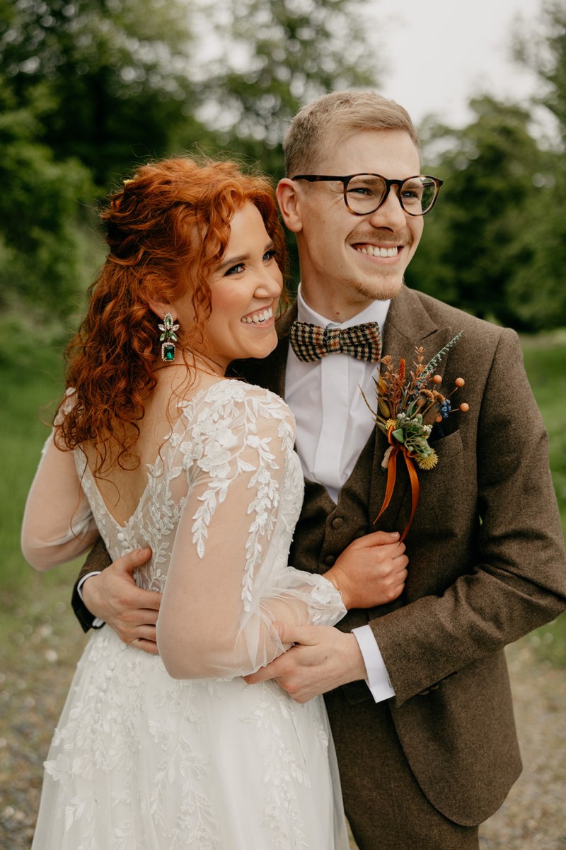 Bride and groom embrace hug in forest trees in background they look away and smile