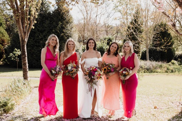 Bride and her bridesmaids stand in the sunshine on the grass posing for a photo