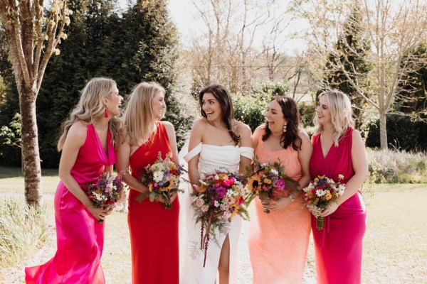 Bride and her bridesmaids stand in the sunshine on the grass posing for a photo