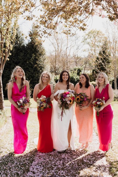 Bride and her bridesmaids stand in the sunshine on the grass posing for a photo