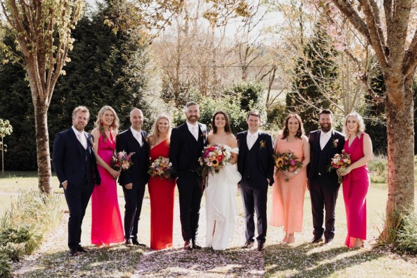 Group portrait photo of bride and groom kissing with bridesmaids and groomsmen on grass in forest