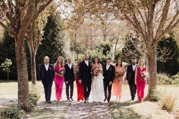 Group portrait photo of bride and groom kissing with bridesmaids and groomsmen on grass in forest