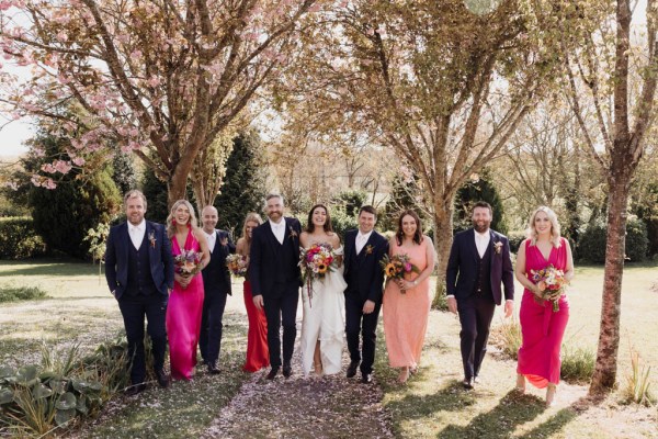 Group portrait photo of bride and groom kissing with bridesmaids and groomsmen on grass in forest