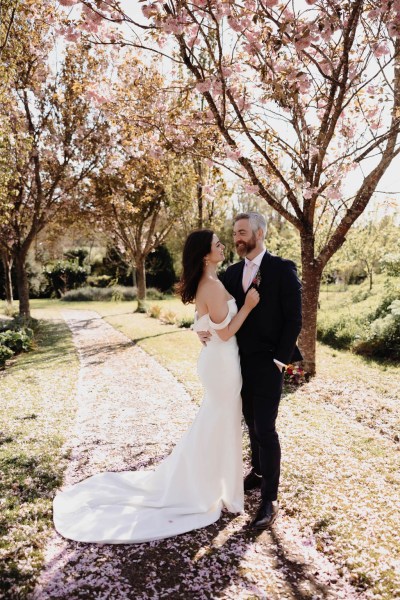 Bride and groom stand in the garden smiling facing each other trees in background