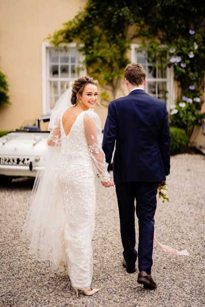 Bride and groom approach white wedding car, bride looks over her shoulder