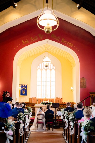 Wide shot of bride and groom seated in front of priest church window