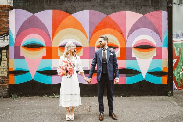 Bride and groom stand holding hands in front of faces painting colourful behind them