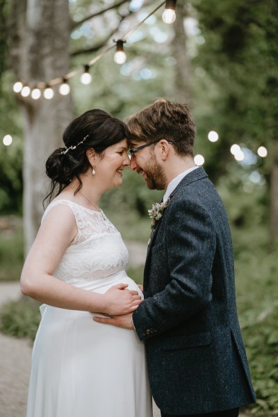 Groom looks at bride as he places hand on her pregnant belly they touch heads